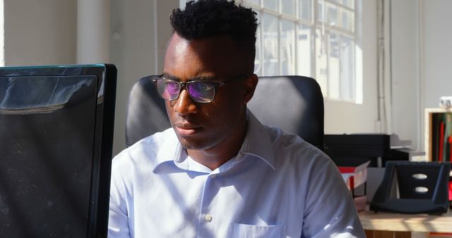 Image depicts a professional man wearing glasses, deeply focused on his computer in a bright, sunlit office space. His serious expression suggests he is diligently working on a task. Ideal for use in business-related articles, corporate websites, and advertisements focused on productivity and work environments.