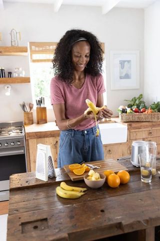 African american woman peeling banana on rustic kitchen island with fresh fruit basket