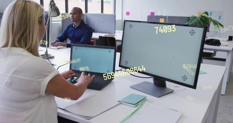 Mature female employee typing on laptop with external monitor in modern open-plan office