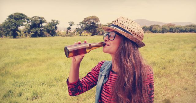 A young Caucasian woman is enjoying a drink from a bottle in a sunny, open field, with copy space. Her casual outfit and straw hat suggest a relaxed day out in nature.