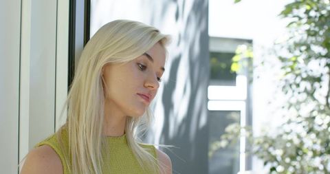 Contemplative blonde woman gazing downward wearing green knit top standing by window