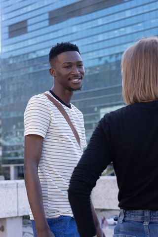 Young man smiling and chatting with friend in urban plaza by modern glass facade
