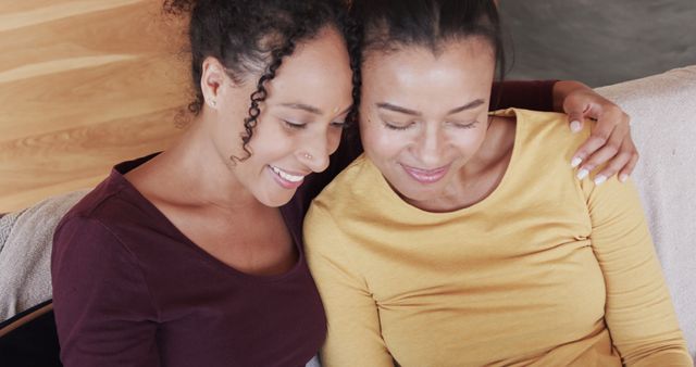 Two women of similar ethnic background are embracing and smiling while sitting indoors. Both are casually dressed, one in a maroon top and the other in a mustard yellow top. They appear content and close, exuding a sense of warmth and togetherness. This image may be used in promotional materials focusing on friendship, family, and emotional well-being. It is also suitable for diverse lifestyle advertisements, blog articles on relationships, or social media posts celebrating friendship.