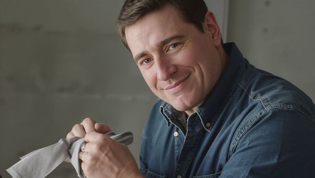 Man polishing metal with cloth, wearing denim shirt, making eye contact