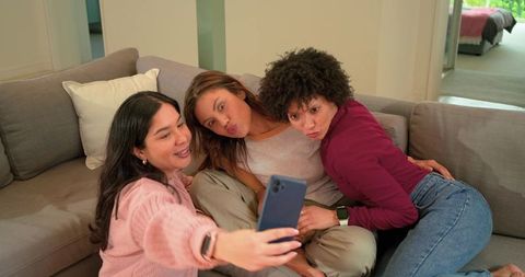 Diverse female friends taking playful selfie on cozy gray sectional sofa at home