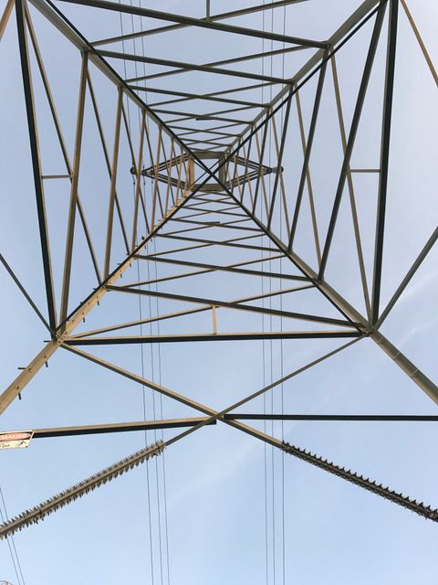 View showing underside of steel electricity transmission tower structure with blue sky in background. Symmetrical lines and geometric shapes are visible creating an abstract, industrial feel. Ideal for use in engineering, architecture, energy industry contexts, or articles about infrastructure and utilities.