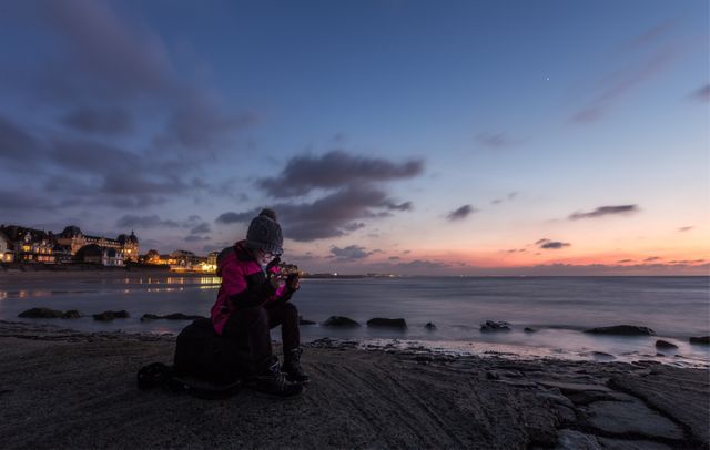 Person sitting on coastal rocks taking a photo of a vibrant sunset with a smartphone. The sky is painted with hues of orange and purple, with scattered clouds. Could be used for travel blogs, technology use in scenic settings, leisure activities, or photography tutorials.