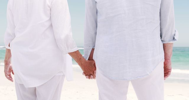 Senior couple holding hands on a beach wearing white clothes. Bright, serene coastal setting enhances themes of togetherness and relaxation. Suitable for illustrating themes of love in old age, retirement, and support in health and wellbeing advertisements.