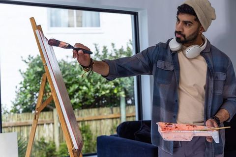 Man painting on easel by window wearing headphones holding palette, brush in sunlit studio
