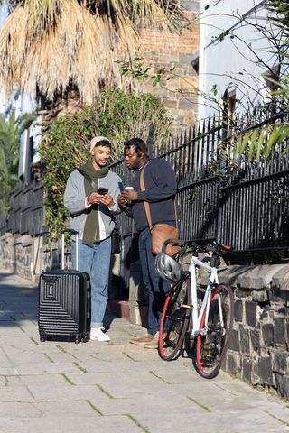 Two friends standing and checking smartphone on urban sidewalk with suitcase and bicycle