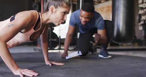 Female athlete doing push-up while male trainer coaching with clipboard in gym