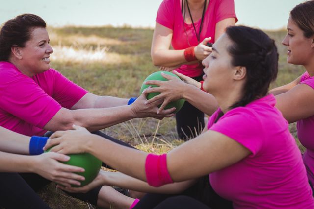 Women participating in an outdoor boot camp under the guidance of a female trainer. They are working in pairs, holding green exercise balls and exchanging them while seated. Ideal for promoting group fitness activities, team building exercises, women's health initiatives, and outdoor workout programs.