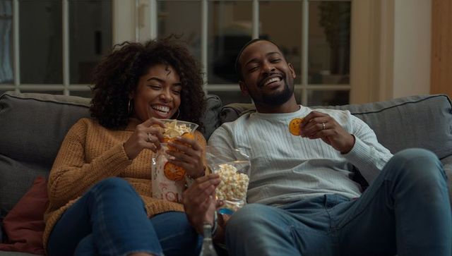 Laughing couple relaxing on gray sofa, sharing popcorn and cookies during cozy night in