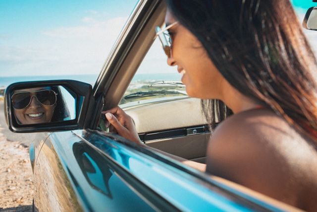 Biracial woman smiling wearing sun glasses looking in wing mirror in convertible car. summer holiday road trip on a country highway by the coast.