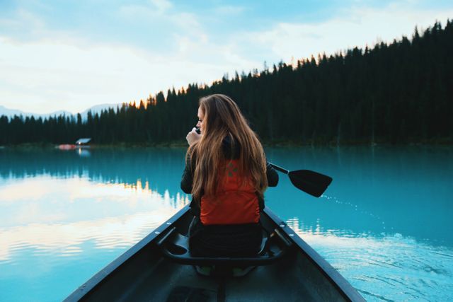 Woman in red life jacket canoeing on calm turquoise mountain lake at sunrise, surrounded by evergreen forest. Perfect for promoting outdoor adventures, nature retreats, and travel destinations. Ideal for use in travel blogs, tourism advertisements, and outdoor equipment promotions.