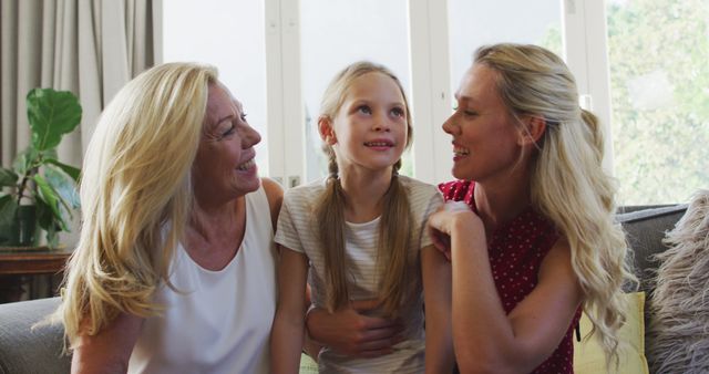 A Caucasian woman, her adult daughter and granddaughter sitting on a couch smiling, the two women kissing the girl. Multi-generation family spending quality time together at home