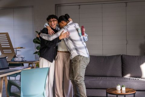Young colleagues sharing warm group hug in modern lounge workspace celebrating teamwork