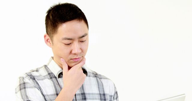 A young Asian man appears contemplative or focused, working on a problem or deep in thought, with copy space. His hand on his chin suggests he's pondering a decision or considering an idea.