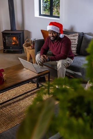 African american man wearing santa hat working on laptop in cozy living room by fireplace