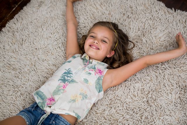 Young girl is happily lying on a soft, textured rug in a living room. She is dressed in casual clothing with a floral top and denim shorts. Her arms are spread out, and she appears to be enjoying a moment of relaxation. This image is perfect for use in advertisements promoting home comfort, family life, or children's products.