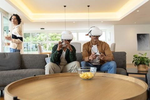 Diverse male teens playing vr games on sofa in sunlit modern living room with snacks and friends