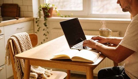 Man working at cozy sunlit kitchen table with laptop, notebook and steaming mug for remote work