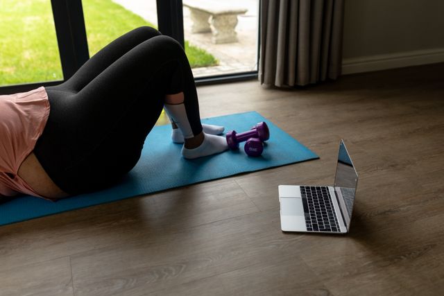 Low section of african american mid adult woman doing exercise while lying on yoga mat by laptop. unaltered, wireless technology, fitness and active lifestyle concept.