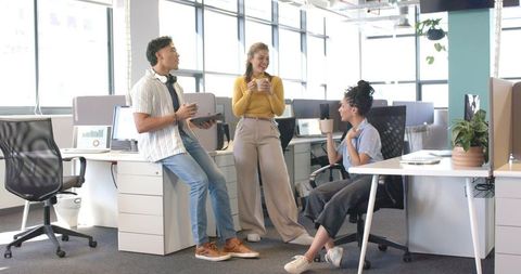 Diverse coworkers chatting over coffee in modern open-plan office, casual teamwork