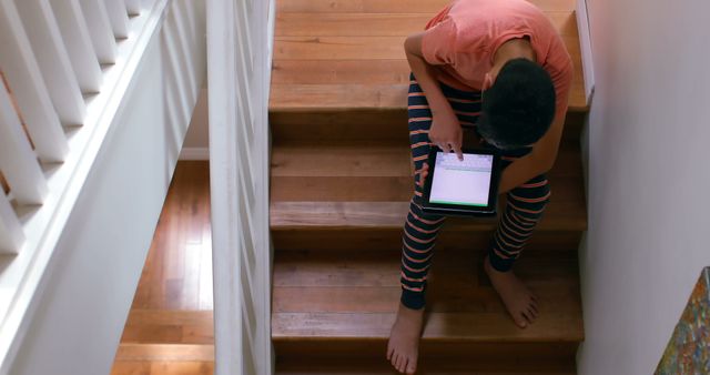 Boy in striped pajamas sitting on wooden stairs, using tablet. Scene highlights modern digital learning environment, casual home interior, and childhood interaction with technology. Useful for illustrating education, digital learning, parenting, and everyday home activities.