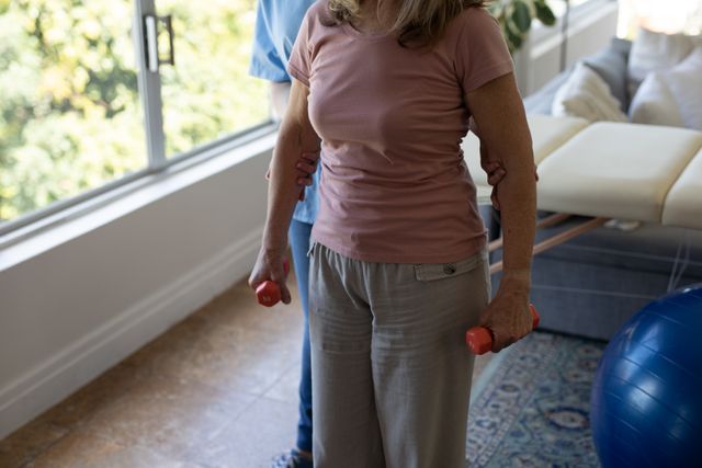 Senior woman exercising with the assistance of a nurse at home, both wearing face masks for safety during the COVID-19 pandemic. Ideal for use in articles or advertisements related to elderly care, home healthcare services, physical therapy, and pandemic safety measures.