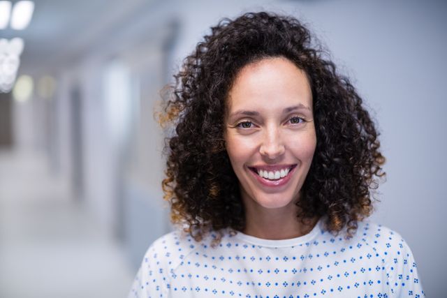 Smiling female patient with curly hair wearing a hospital gown in a hospital corridor. Ideal for use in healthcare, medical, and wellness contexts, including patient care, recovery, and hospital environments.