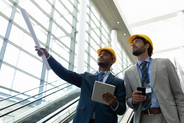 Two male architects in business attire and hard hats are discussing construction plans in a modern office building. One is holding blueprints while the other is using a tablet. This image is ideal for illustrating concepts related to architecture, engineering, project management, teamwork, and modern office environments.