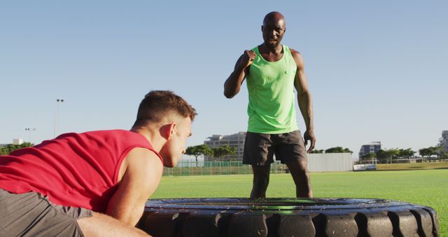 Fitness coach giving instructions to a man performing a tire flip exercise on a sunny day. Suitable for articles or advertisements on outdoor fitness, guided workouts, athletic training, and exercise routines.
