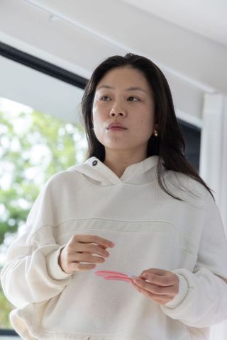 Asian woman holding pink plastic pieces in white hoodie with gold hoops, contemplative natural light