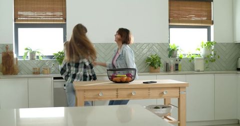 Sunlit kitchen dance: two friends laughing and dancing around wooden island with fruit bowl