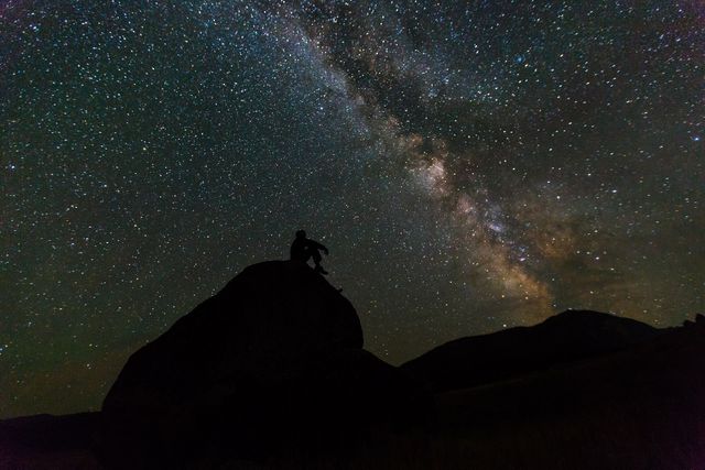 Silhouette of a person sitting on a rock, engulfed in the brilliant display of the Milky Way and countless stars in the night sky. The scene evokes a sense of serenity and wonder, perfect for themes of astronomy, night-time adventures, and natural beauty. Ideal for use in astronomy blogs, travel magazines, outdoor adventure advertisements, and inspirational posters.