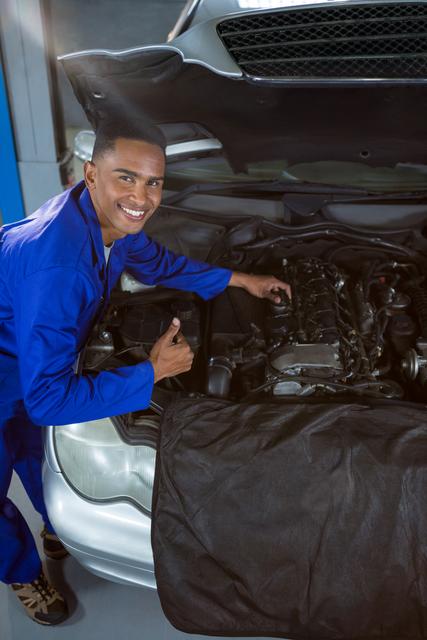 Mechanic in blue overalls smiling and giving thumbs up while working on car engine in garage. Ideal for use in automotive service advertisements, mechanic training materials, and promotional content for car repair shops.