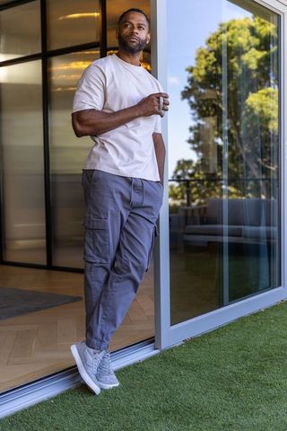 African american mature man leaning on sliding glass door holding mug, modern home living