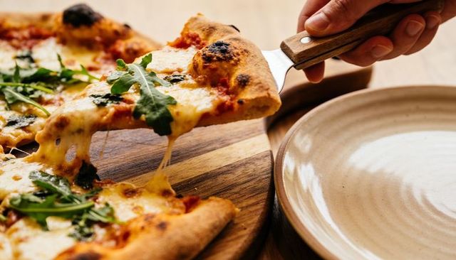 Lifting wood-fired pizza slice with melting mozzarella and fresh arugula on wooden board