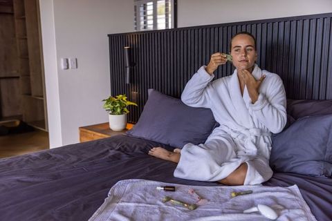 Woman using jade roller on face in white robe on bed for morning skincare routine