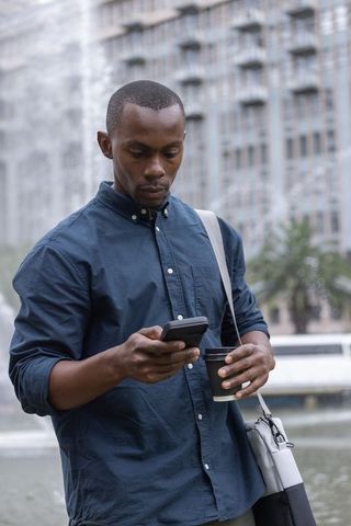 African american commuter checking phone while holding coffee at urban plaza fountain