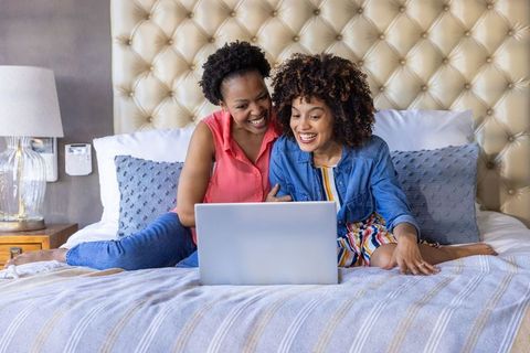Diverse female couple relaxing on bed using laptop, sharing joyful moment