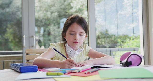 Caucasian girl in face mask on chin sitting on table and writing in notebook. Education, learning, health, hygiene, coronavirus, childhood and domestic life, unaltered.