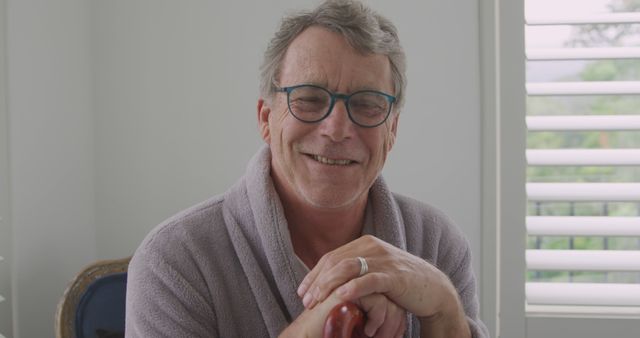 Senior man, wearing glasses and a purple bathrobe, smiling relaxedly while sitting at home. Ideal for illustrating themes of retirement, comfort, home life, casual moments, and senior lifestyle.