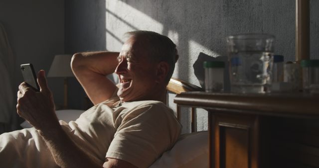 Senior man lying in bed while smiling and using a smartphone. Morning light illuminates the room. The man looks relaxed and happy as he interacts with technology. Use this image for promoting products or services related to seniors, technology use among elderly, relaxation, and morning routines.