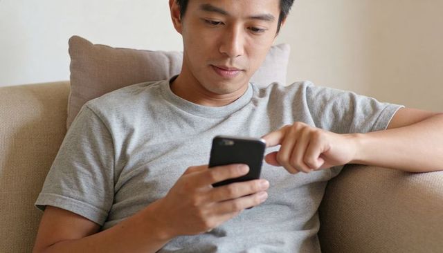 Asian man scrolling smartphone on sofa, relaxed minimalist home lifestyle moment