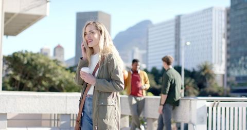 Smiling woman holding coffee and smartphone on sunlit urban terrace with friends nearby