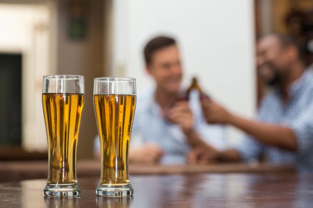 Two tall beer glasses filled with golden beer on a wooden table in a restaurant. In the background, two men are clinking bottles, suggesting a social and celebratory atmosphere. Ideal for use in advertisements for bars, pubs, restaurants, or social events. Can also be used in articles about socializing, beer culture, or relaxation.