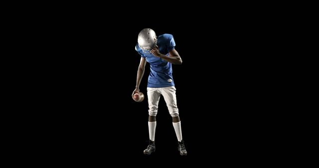An African American teenage boy in a football uniform holds his helmet under his arm, with copy space. His posture suggests a moment of contemplation or defeat after a game or during a break in play.