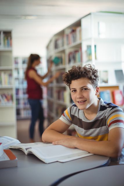 Young boy with curly hair smiling while studying in a library. Ideal for educational content, school promotions, academic articles, and learning resources. Background shows bookshelves and another person searching for a book, emphasizing a studious environment.